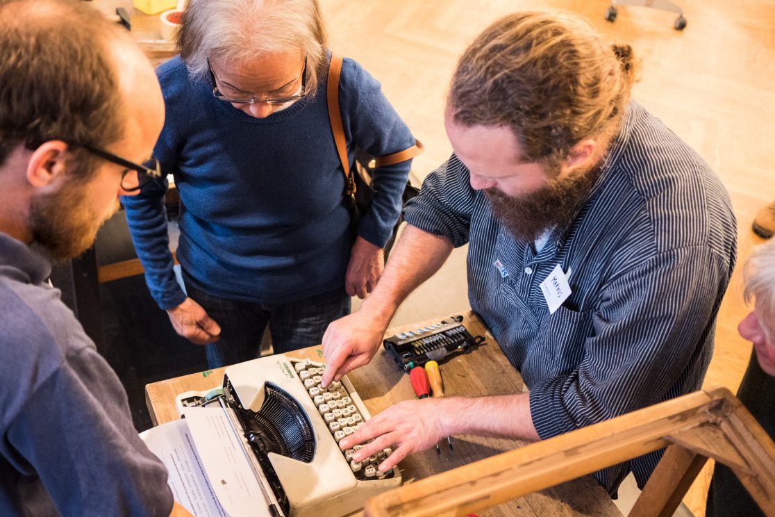 Foto: Drei Männer stehen um einen Tisch herum und schauen auf eine Schreibmaschine, einer der drei Männer tippt auf dieser