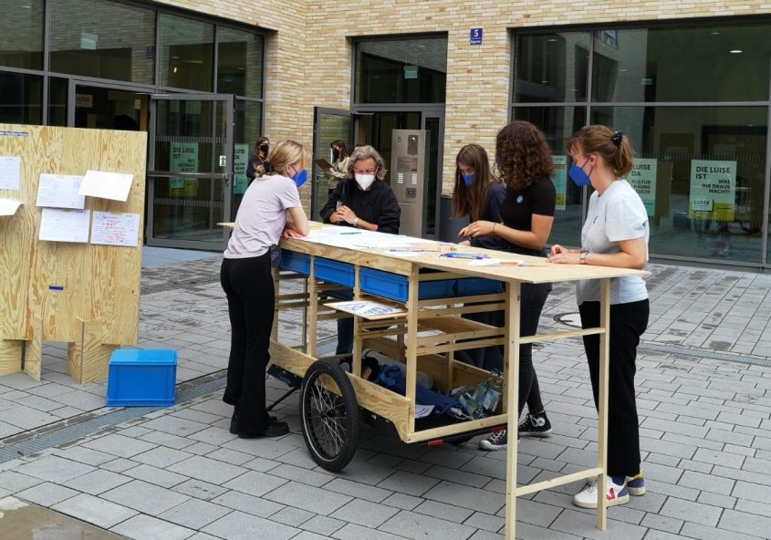 An einem umgebauten Fahrradanhänger unterhalten sich mehrere Menschen und zeigen auf Materialien.