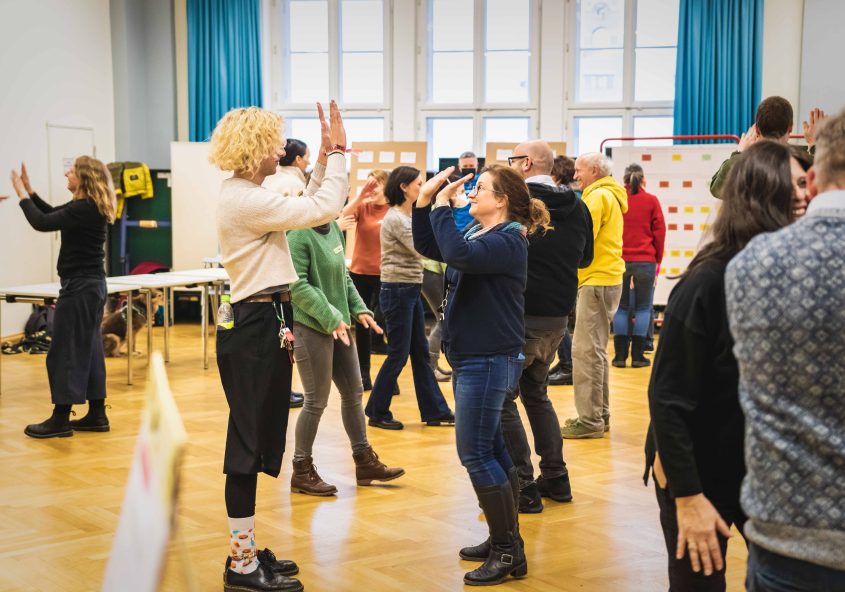 zwei Frauen im Zentrum stehen sich gegenüber und klatschen sich in die Hände, umgeben von anderen Teilnehmer:innen