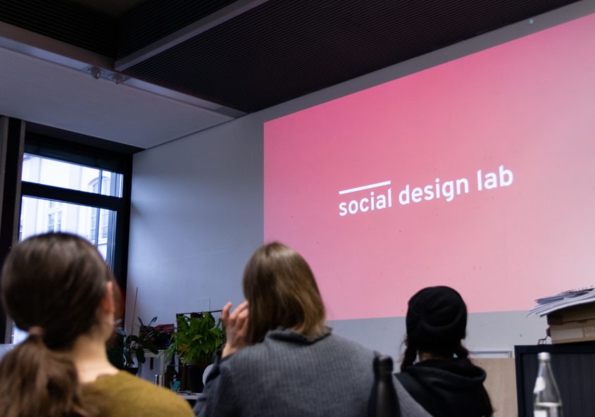 Studierende schauen auf eine Projektionsfläche auf der das Logo des social design labs zu sehen ist.