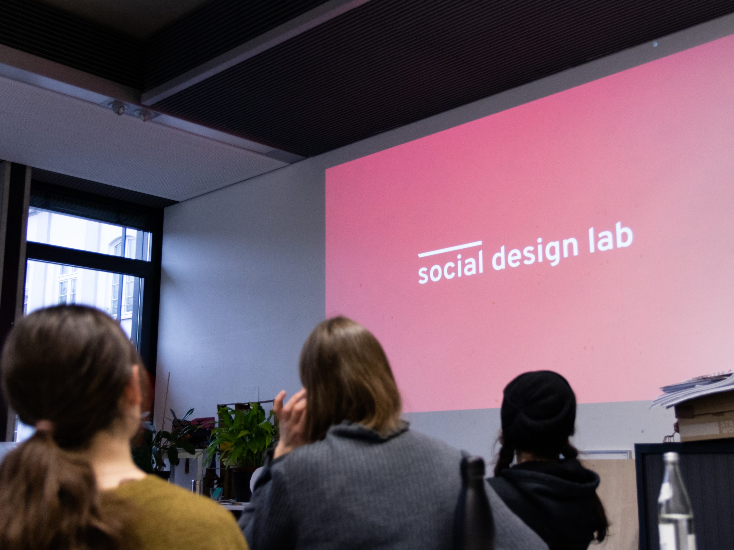 Studierende schauen auf eine Projektionsfläche auf der das Logo des social design labs zu sehen ist.