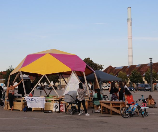 ein aus Holz und Planen konstruierter Bau, der an ein Zelt erinnert, steht auf einem großen asphaltierten Platz. In dem Bau sind Familien und spielen, malen und unterhalten sich.