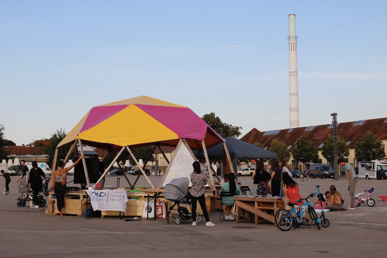 Habibi22_9741 3 ein aus Holz und Planen konstruierter Bau, der an ein Zelt erinnert, steht auf einem großen asphaltierten Platz. In dem Bau sind Familien und spielen, malen und unterhalten sich.