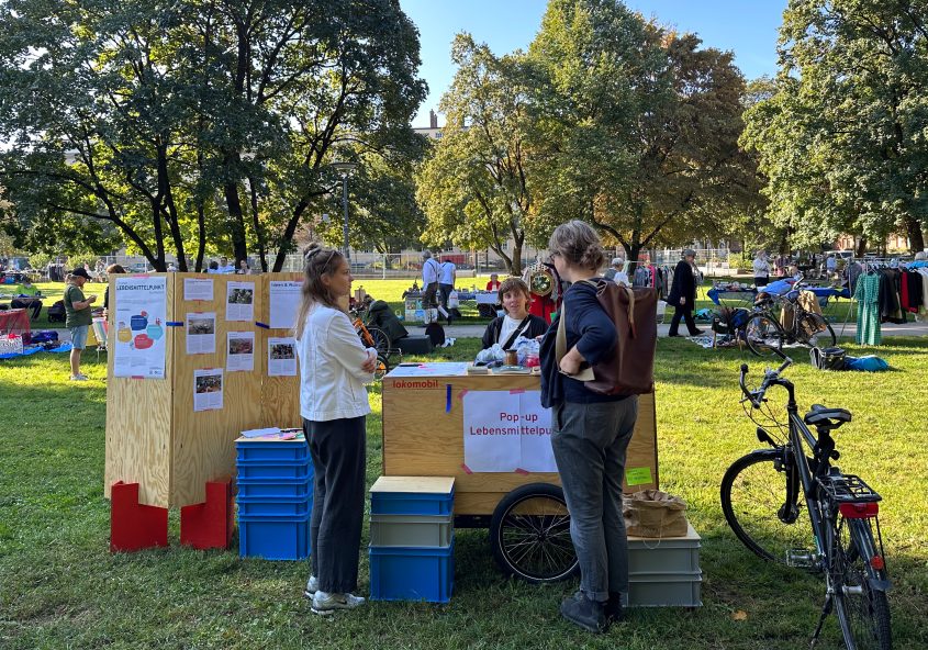 Mitarbeiterinnen des social design lab mit einem Pop-up Stand bei einer Veranstaltung in München während einem Flohmarkt in einem Park, sprechen mit einer Frau