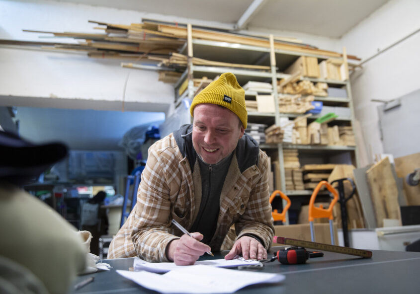 Eine lächelnde Person des treibgut-Teams schreibt, über einen Schreibtisch gebeugt, etwas in ein Dokument.