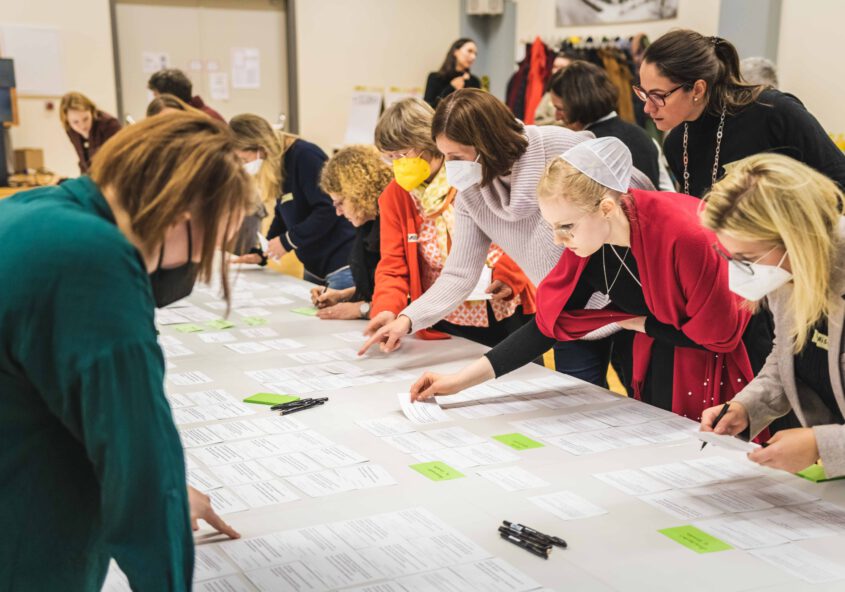 Eine Gruppe von Menschen, meist Frauen, ist an einem langen Tisch mit Papierblättern und Notizen, einige davon grün markiert, in eine gemeinschaftliche Aktivität eingebunden. Die Teilnehmer sind leger gekleidet, tragen FFP2-Masken und scheinen konzentriert zu sein, während sie auf die Zettel zeigen und darauf schreiben. Die Umgebung wirkt wie eine Gemeinde oder ein Bildungszentrum, mit einem hellen Raum und einigen Gegenständen, die im Hintergrund hängen.