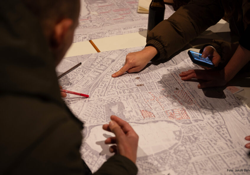 Menschen sind um einen Tisch versammelt und schauen auf eine große Karte. Es sind Hände und eine Person von hinten zu sehen. Eine Person zeigt auf die Karte, während andere Stifte und Smartphones halten.