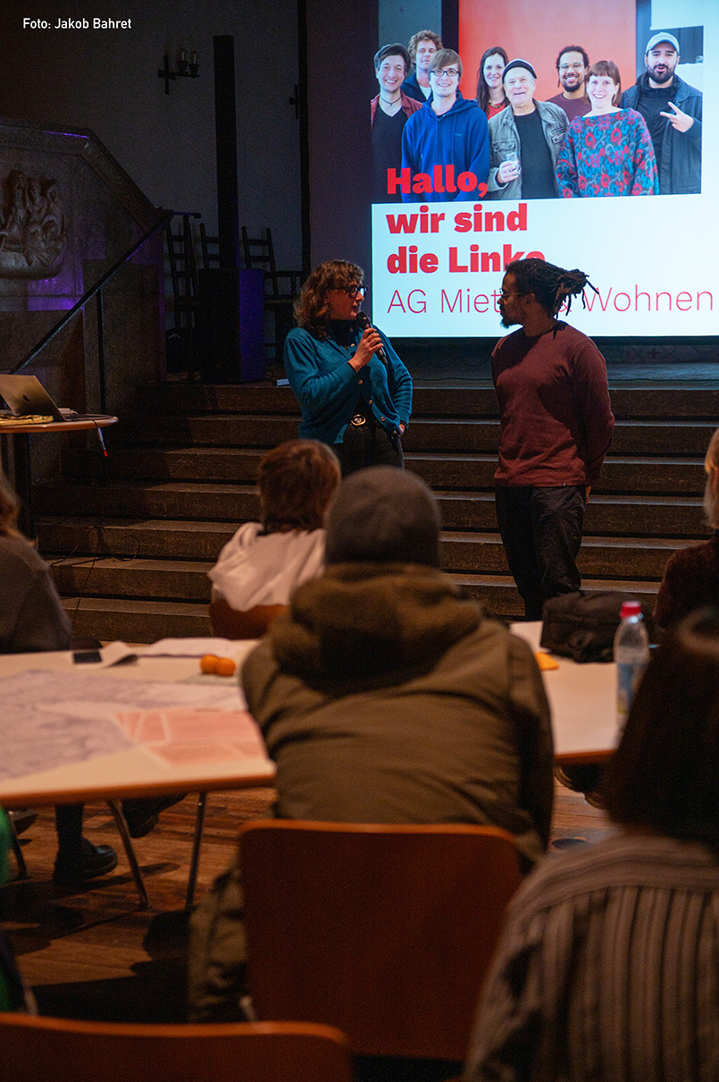 Eine Frau mit lockigem Haar und Brille spricht in ein Mikrofon, während ein Mann mit Dreadlocks aufmerksam zuhört.Sie stehen auf der Bühne vor einem großen Bildschirm, auf dem der Text „Hallo, wir sind die Linke AG Mieten-Wohnen“ und Gruppenfotos von mehreren Personen zu sehen sind.Im Vordergrund sitzt ein bunt gemischtes Publikum an Tischen mit Papieren und Wasserflaschen und konzentriert sich auf die Präsentation. Die Personen sind in einer Kirche. Diese ist sanft beleuchtet, was die intime Atmosphäre der Veranstaltung unterstreicht.