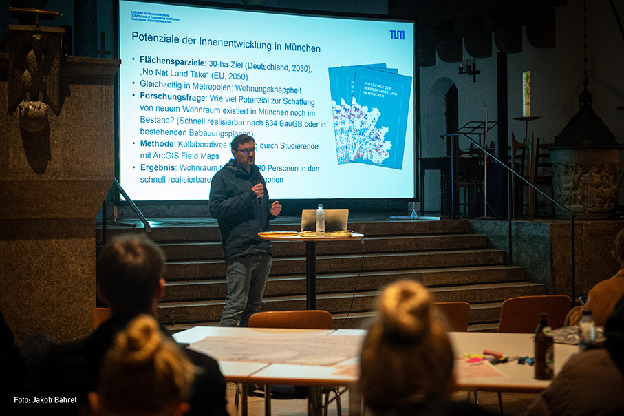 Ein Mann steht an einem Podium und spricht zu einem Publikum in einem schwach beleuchteten Raum. Hinter ihm wird auf einer großen Projektionsfläche eine Präsentation über die Stadtentwicklung in München gezeigt. Auf der Leinwand sind Aufzählungspunkte auf Deutsch zu sehen, die Themen wie Flächennutzung, Wohnungsnot und Forschungsmethoden behandeln. Mehrere blaue Broschüren mit dem Titel Potenziale der Innenentwicklung in München sind auf der Leinwand zu sehen. Die Zuhörer, die an Tischen sitzen, sehen engagiert aus, einige machen sich Notizen, andere schauen aufmerksam zu. Auf dem Tisch vor dem Referenten stehen eine Wasserflasche und Papiere.