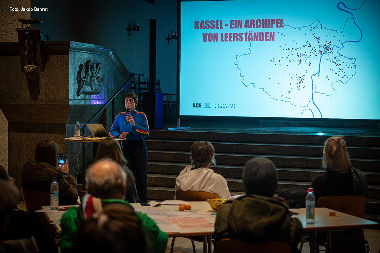 Eine Person steht auf einem Podium und spricht zu den Zuhörern, während hinter ihr auf einem großen Bildschirm eine Karte mit der Überschrift KASSSEL - EIN ARCHIPEL VON LEERSTÄNDEN zu sehen ist. Auf der Karte sind verschiedene Orte mit Punkten markiert, die auf leere Flächen in der Stadt hinweisen. Die Person, gekleidet in einen bunten Pullover, hält ein Mikrofon in der Hand und scheint mit dem Publikum, das an Tischen mit Papieren und persönlichen Gegenständen sitzt, in Kontakt zu treten. Die Kulisse besteht aus Steinwänden, gedämpfter Beleuchtung und dekorativen Elementen, die eine akademische Atmosphäre schaffen.