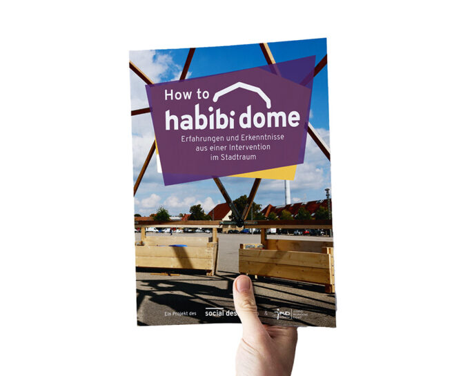 Ein Foto, auf dem eine Hand zu sehen ist, die das Manual „How to Habibi Dome“ vor einem weißen Hintergrund hält.