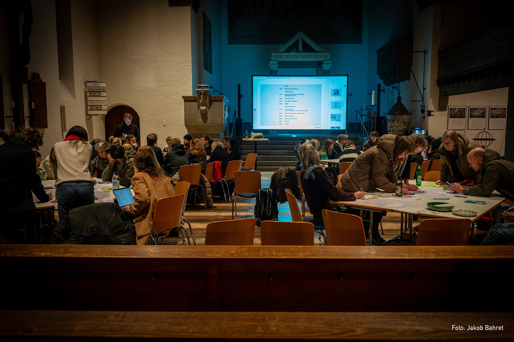 Verschiedene Personengruppen arbeiten an großen Tischen in einem Kirchenraum an diversen Karten und anderen Aufzeichnungen. Hinter den Tischen ist ein großer Screen mit einem Präsentations-Slide zu sehen.