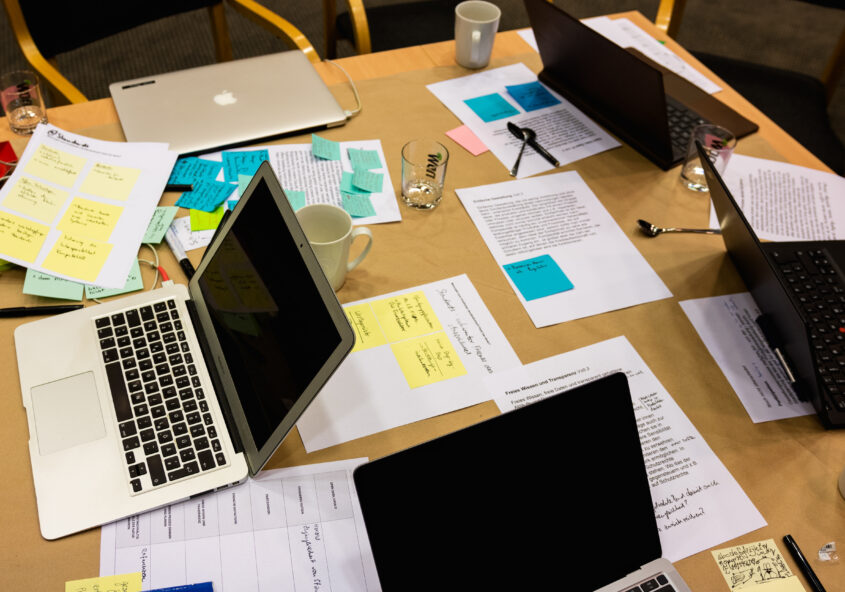 Ein Schreibtisch des Circular Society Kollegs, schräg von oben fotografiert. Zu sehen sind mehrere Laptops sowie ausgedruckte Unterlagen, Post-its, Stifte und Wassergläser. Das Foto macht deutlich, dass hier eine Gruppe intensiv an etwas arbeitet oder forscht.