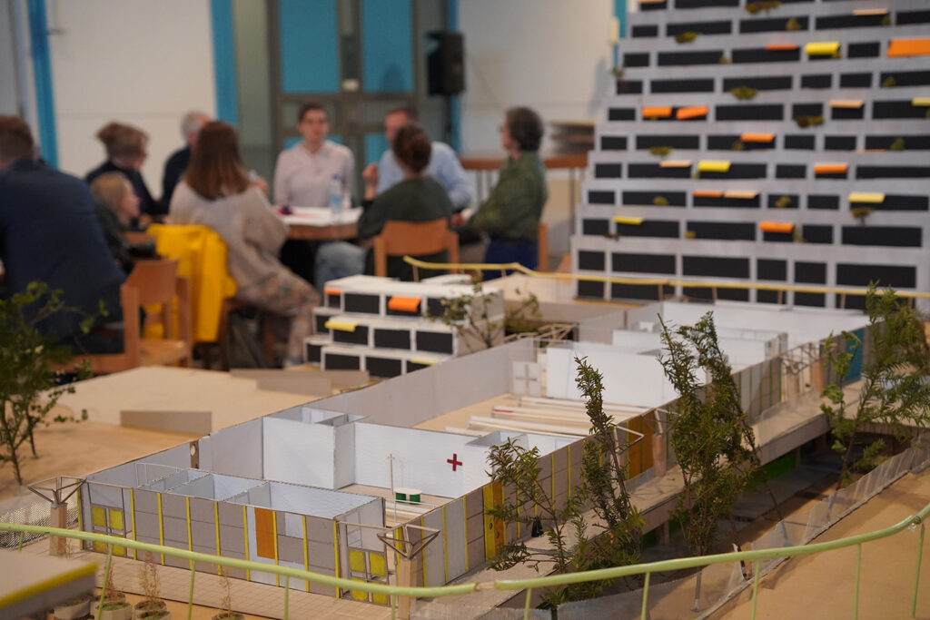 Das Bild zeigt ein Architekturmodell, das im Vordergrund steht und einen Gebäudekomplex mit mehreren kleinen Gebäuden und Grünflächen darstellt. Im Hintergrund ist eine unscharf dargestellte Gruppe von Personen zu sehen, die an einem Tisch sitzt und sich zu unterhalten scheint. Es handelt sich wahrscheinlich um eine Präsentation oder ein Workshop zu einem Architekturprojekt. Im Hintergrund ist ein weiteres, größeres Architekturmodell zu erkennen - ein Hochhaus mit vielen kleinen, farbig markierten Balkonen.