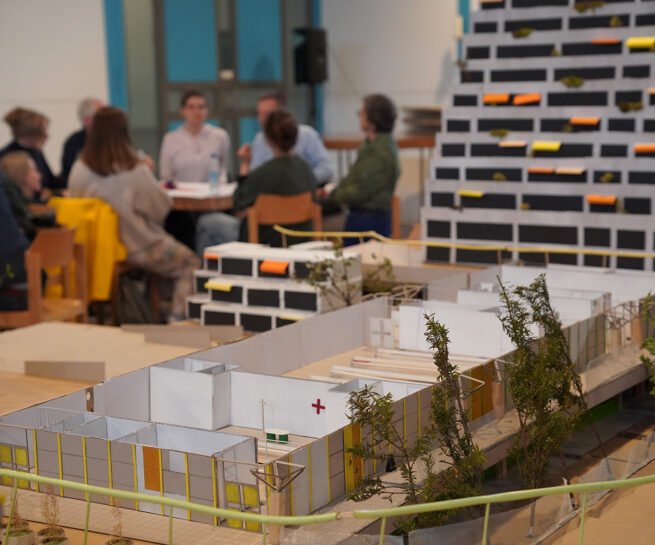 Das Bild zeigt ein Architekturmodell, das im Vordergrund steht und einen Gebäudekomplex mit mehreren kleinen Gebäuden und Grünflächen darstellt. Im Hintergrund ist eine unscharf dargestellte Gruppe von Personen zu sehen, die an einem Tisch sitzt und sich zu unterhalten scheint. Es handelt sich wahrscheinlich um eine Präsentation oder ein Workshop zu einem Architekturprojekt. Im Hintergrund ist ein weiteres, größeres Architekturmodell zu erkennen - ein Hochhaus mit vielen kleinen, farbig markierten Balkonen.