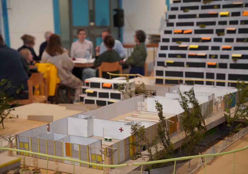 Das Bild zeigt ein Architekturmodell, das im Vordergrund steht und einen Gebäudekomplex mit mehreren kleinen Gebäuden und Grünflächen darstellt. Im Hintergrund ist eine unscharf dargestellte Gruppe von Personen zu sehen, die an einem Tisch sitzt und sich zu unterhalten scheint. Es handelt sich wahrscheinlich um eine Präsentation oder ein Workshop zu einem Architekturprojekt. Im Hintergrund ist ein weiteres, größeres Architekturmodell zu erkennen - ein Hochhaus mit vielen kleinen, farbig markierten Balkonen.