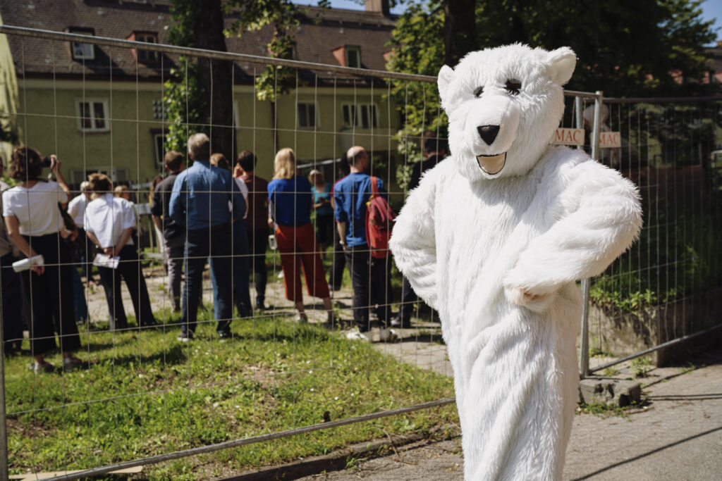 Eine Person in einem Eisbärkostüm steht vor einer Gruppe von Menschen, die auf ein anscheinend städtisches Gebäude blickt. Das Foto entstand während einer Bustour des social design lab und dessen Partnerorganisationen.
