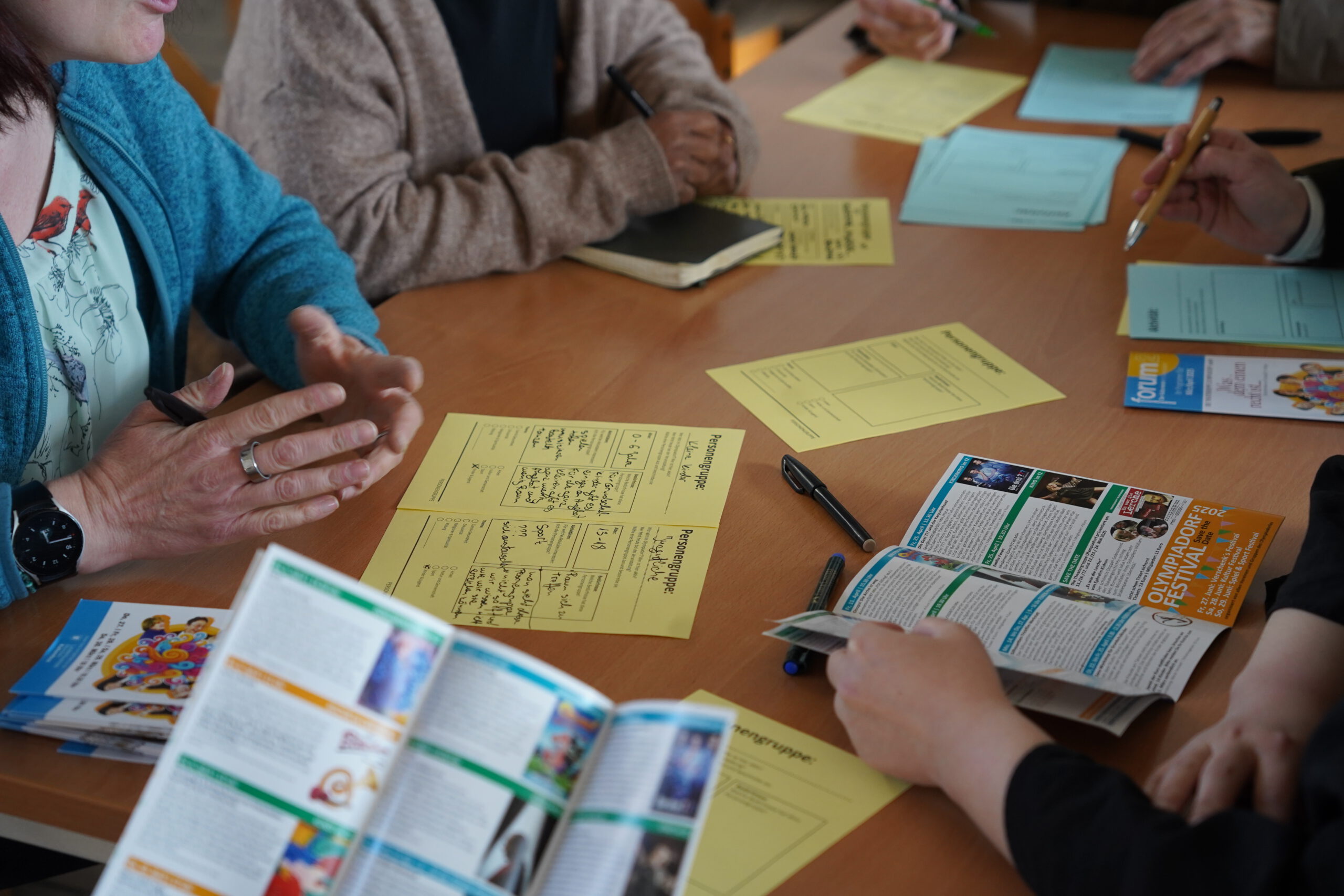 Ein Tisch mit verschiedenen Materialien, unter anderem ausgefüllten Vorlagen und Informationsbroschüren, an dem verschiedene Menschen sitzen und miteinander im Gespräch sind.