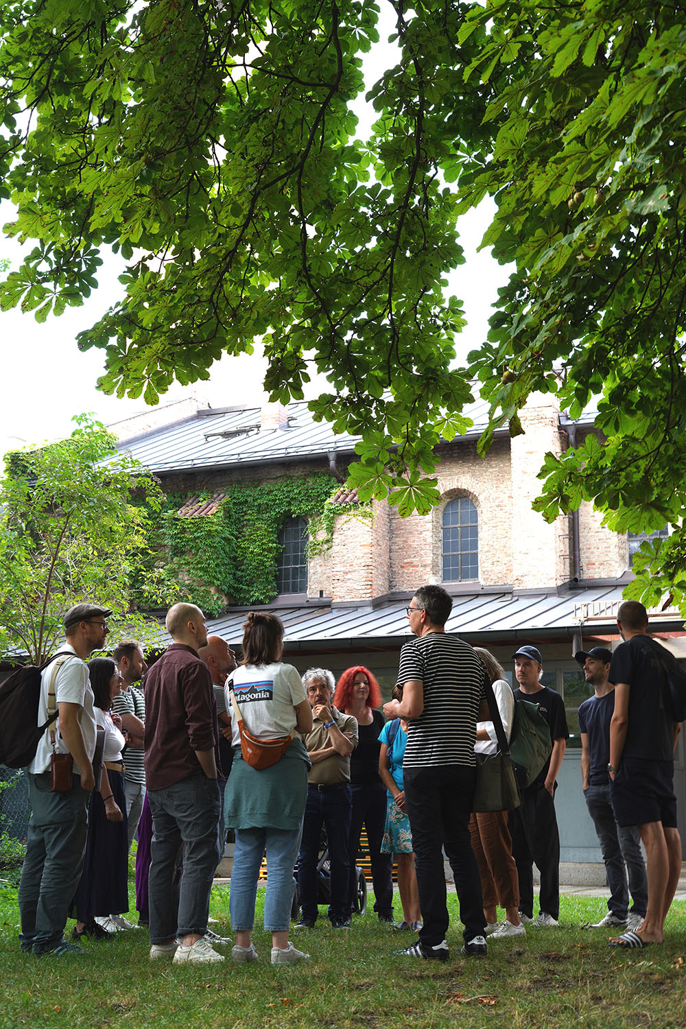 Das Bild zeigt eine Gruppe von etwa 15 Personen, die sich unter einem Baum auf einer Rasenfläche vor einem alten Backsteingebäude versammelt haben. Die Gruppe scheint aufmerksam einem Mann zuzuhören, der im Zentrum steht und etwas zu erklären scheint – das Bild entstand im Rahmen einer der Kartierungsspaziergänge des social design lab und ihrer Partner*innen in München. Das Gebäude im Hintergrund wirkt historisch und ist teilweise von Efeu bedeckt. Die Atmosphäre ist entspannt und eher informell.