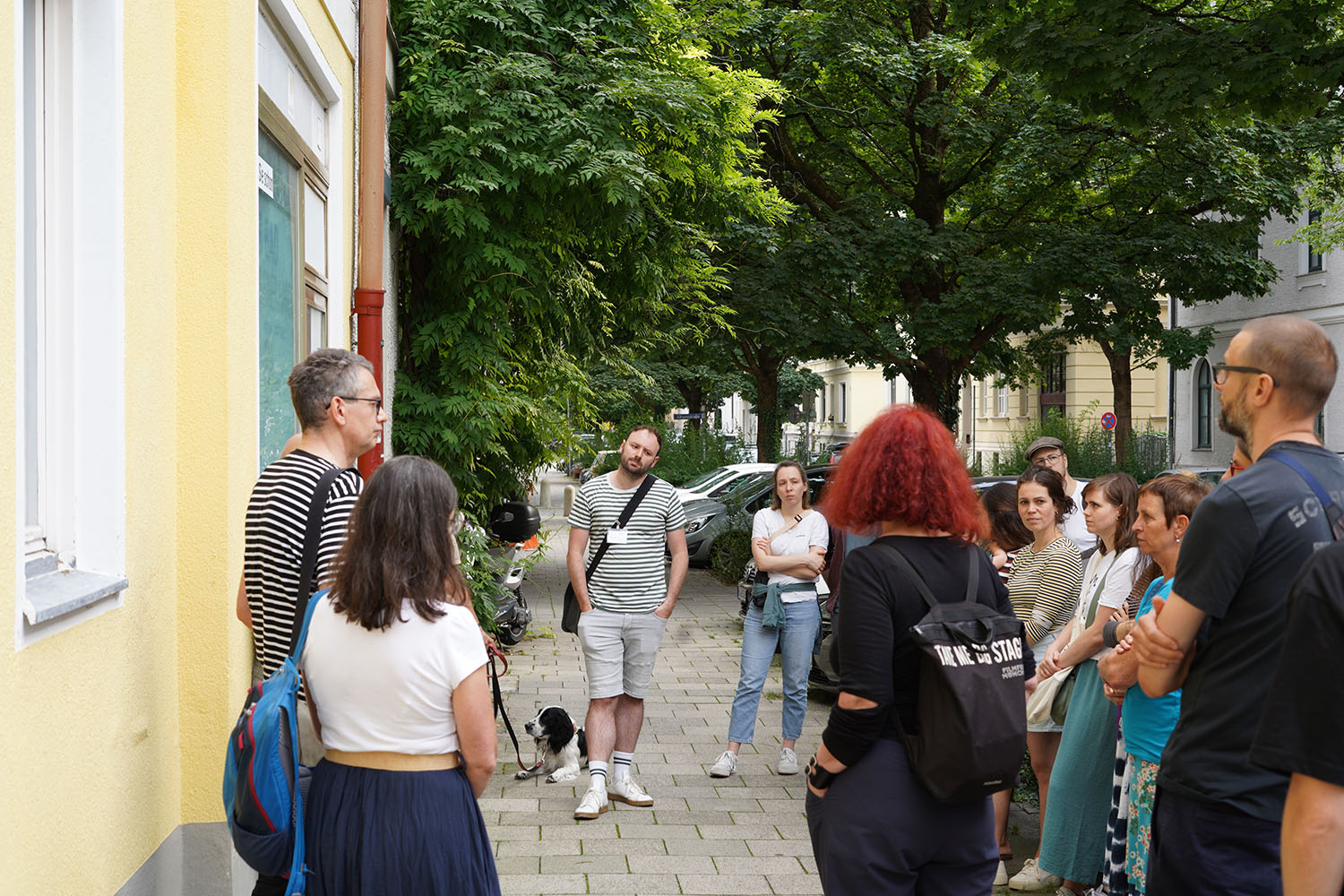 Mehrere Personen stehen im Kreis auf der Straße in einer Münchner Nachbarschaft und hören einer sprechenden Person zu.