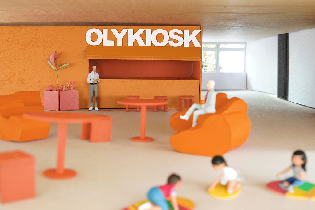 Ein Miniaturmodell eines lebendigen Innenraums mit verschiedenen Orangetönen. Im Mittelpunkt steht ein Kiosk mit der Aufschrift „OLYKIOSK“, hinter dem eine Person steht. Im Vordergrund befinden sich farbenfrohe Loungemöbel, darunter ein großer orangefarbener Sessel und ein kleiner Tisch. Kinder spielen auf runden Matten auf dem Boden und sorgen für eine lebhafte Atmosphäre. Einige dekorative Pflanzen unterstreichen die Wärme und Einladendheit des Designs. Große Fenster lassen natürliches Licht herein und erhellen den Raum.