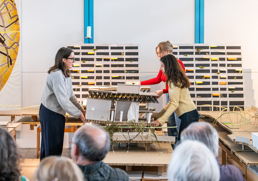 Das Bild zeigt drei junge Frauen, die konzentriert an einem Architekturmodell arbeiten. Sie scheinen es gemeinsam zu bewegen oder zu positionieren. Das Modell ist aus Pappe gefertigt und zeigt ein Gebäude mit viel Grün. Im Hintergrund sind weitere Modelle und eine Wand mit kleinen Kärtchen zu sehen, was auf eine Präsentation oder einen Workshop hindeutet. Im Vordergrund sitzen Zuschauer, die der Präsentation folgen.