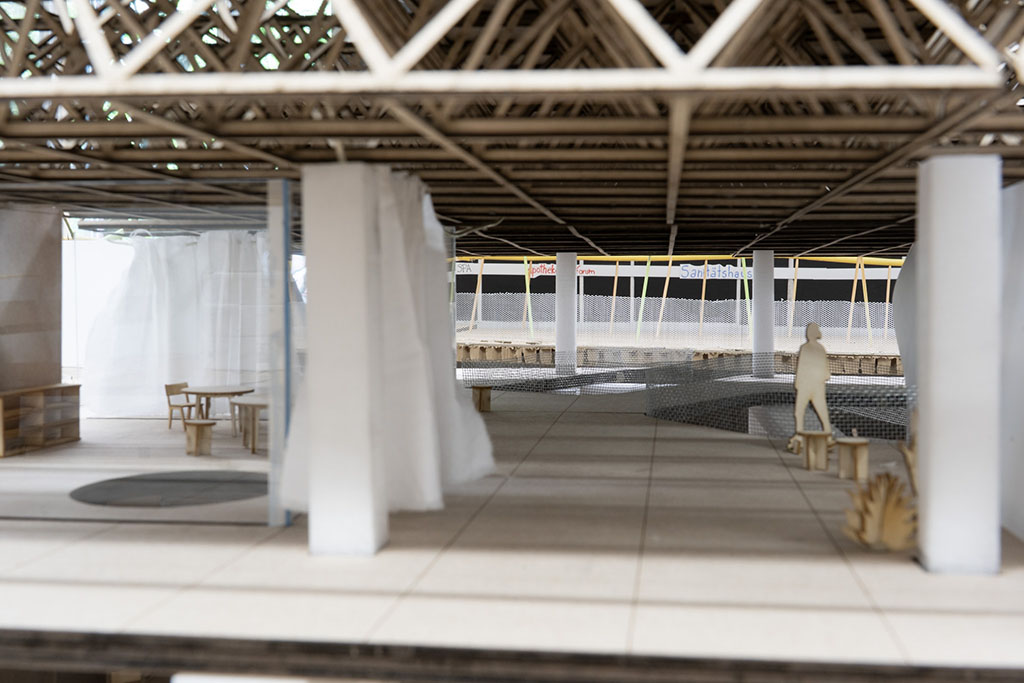 Ein detailliertes Architekturmodell eines geräumigen, offenen Innenraums mit einer komplexen, gitterartigen Decke. Der Boden besteht aus hellen Fliesen, dazu kommen minimalistische Holzmöbel, darunter ein kleiner Tisch und Stühle. Ein Bereich ist teilweise durch transparente weiße Stoffvorhänge abgetrennt, was ein Gefühl von Privatsphäre vermittelt. Die Silhouette einer menschlichen Figur steht in der Nähe einer Bank und vermittelt einen Eindruck von der Größe des Raums. Der Hintergrund enthält Andeutungen von Außenelementen, darunter Plakate mit Namen oder Orten, die den Kontext und die Lebendigkeit des Modells unterstreichen.