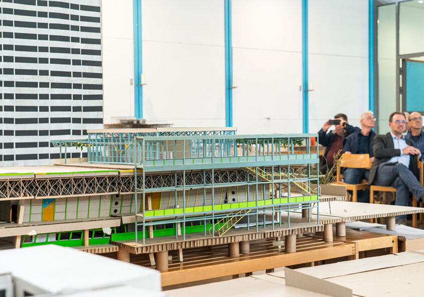 Das Bild zeigt ein Architekturmodell eines mehrstöckigen Gebäudes, das auf einer Plattform aus Pappe ruht. Das Modell ist detailliert und zeigt die Struktur des Gebäudes mit Treppen, Balkonen und einer begrünten Dachterrasse. Im Hintergrund sind mehrere Personen zu sehen, die dem Modell zu folgen scheinen.