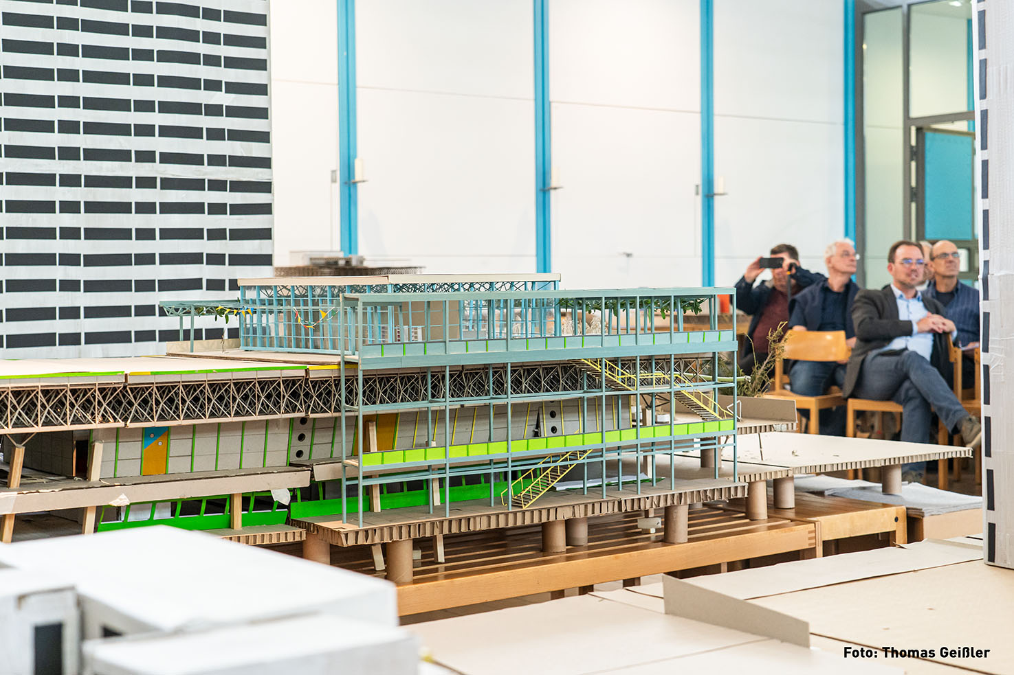 Das Bild zeigt ein Architekturmodell eines mehrstöckigen Gebäudes, das auf einer Plattform aus Pappe ruht. Das Modell ist detailliert und zeigt die Struktur des Gebäudes mit Treppen, Balkonen und einer begrünten Dachterrasse. Im Hintergrund sind mehrere Personen zu sehen, die dem Modell zu folgen scheinen.