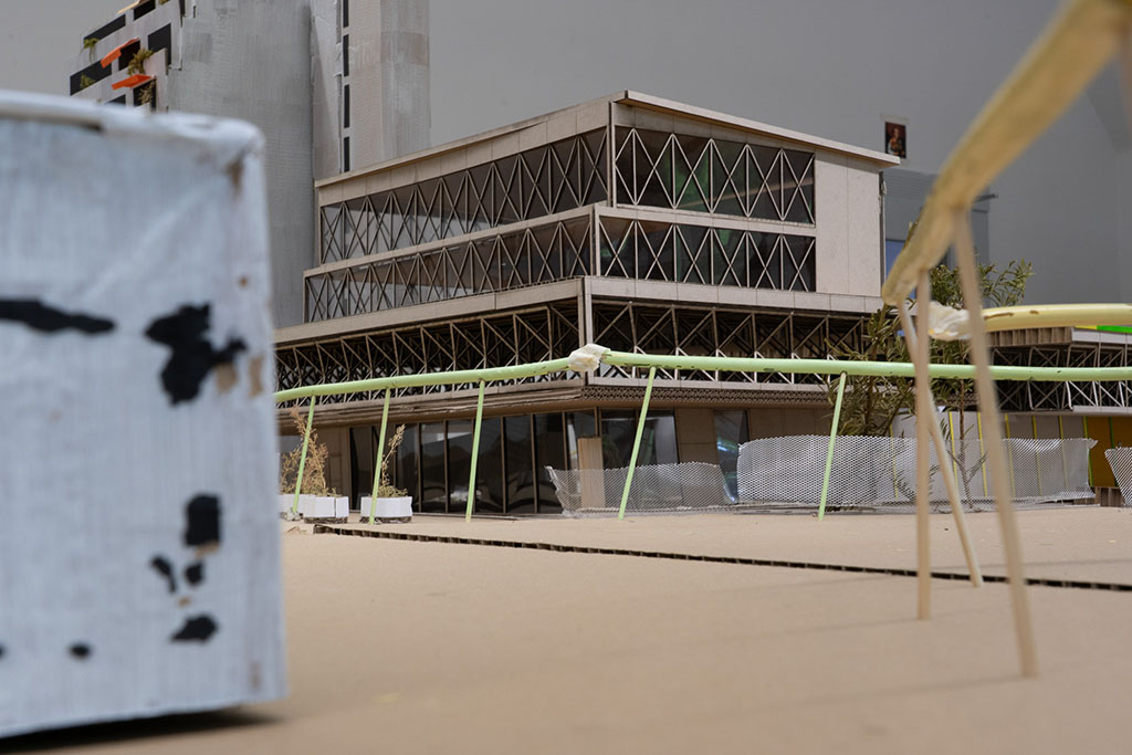 Ein detailliertes Architekturmodell zeigt ein mehrstöckiges Gebäude mit modernem Design, das sich durch eine Kombination aus Glas und strukturierten Oberflächen auszeichnet. Die Fassade weist komplexe Rautenmuster auf, und es gibt einen Balkon mit grünen Geländern. Im Vordergrund ist eine strukturierte Kartonoberfläche zu sehen, während der Hintergrund eine einfache Bauumgebung mit gedeckten Farben und Materialien zeigt, was auf ein unfertiges Projekt hindeutet. Kleine Pflanzen und Schutzgitter verleihen der Modellkulisse zusätzliche Realitätsnähe.