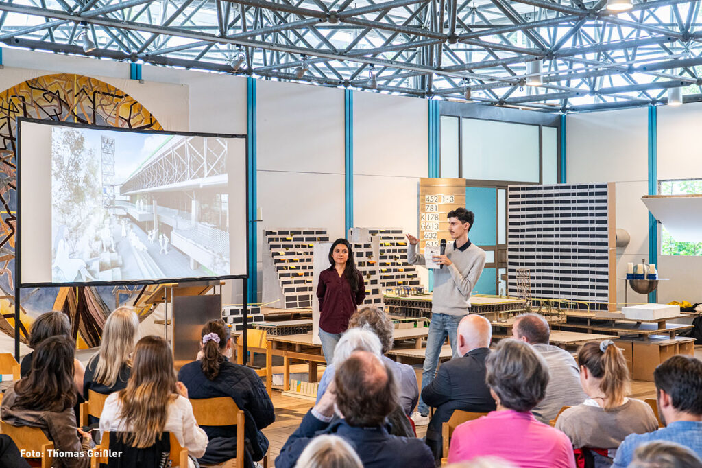 Das Bild zeigt eine Präsentation vor einem Publikum. Zwei Personen stehen vor einem Architekturmodell und einem Projektor, auf dem eine Architekturzeichnung zu sehen ist. Eine Person hält ein Mikrofon und präsentiert anscheinend das Projekt. Das Publikum sitzt auf Stühlen und verfolgt aufmerksam die Präsentation. Der Raum wirkt wie eine Ausstellungshalle oder ein Hörsaal, es handelt sich um einen Raum des Ökumenischen Kirchenzentrums im Olympiadorf München.