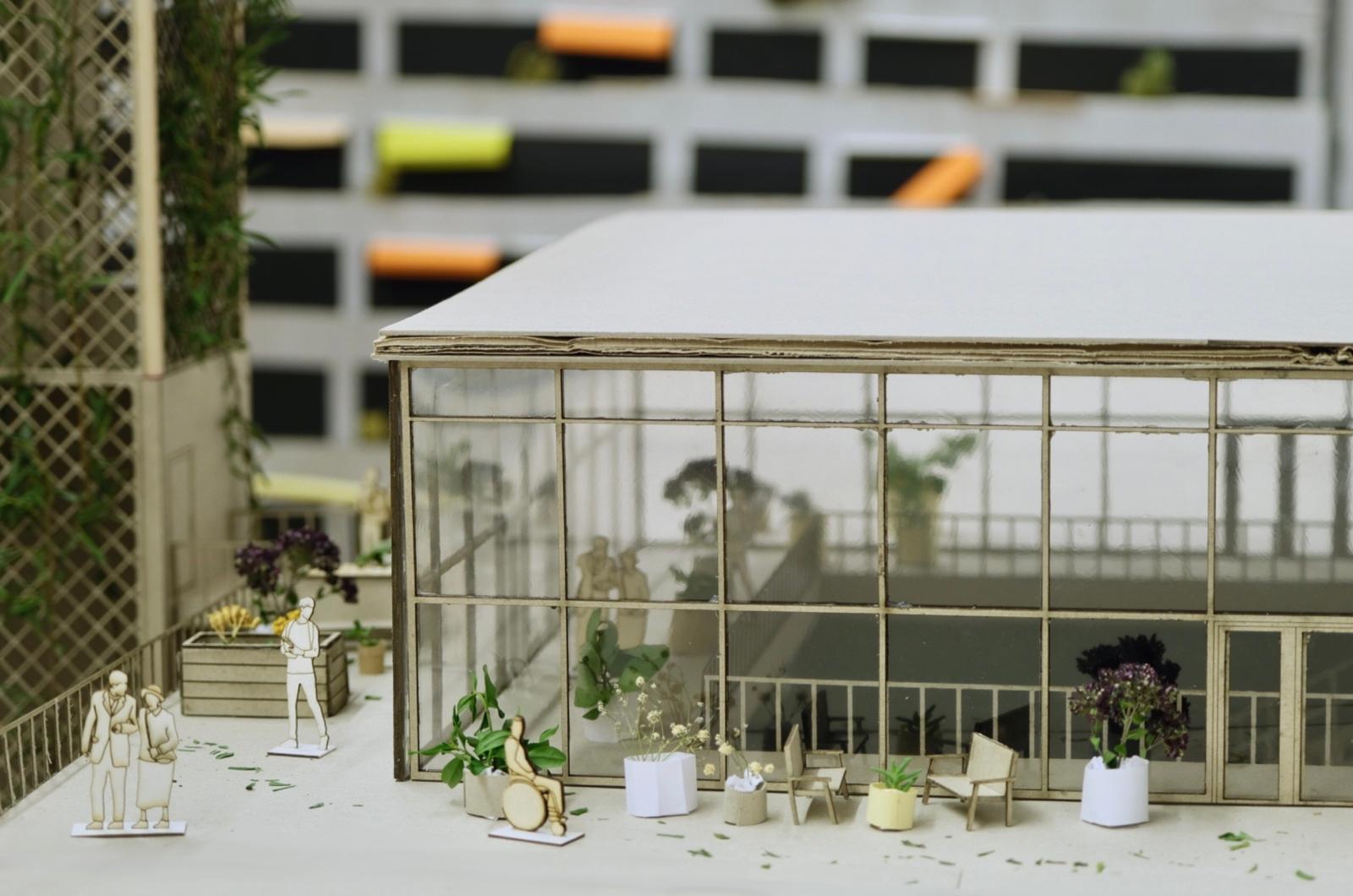 Ein detailliertes Architekturmodell eines modernen Gebäudes mit Glasfassade, umgeben von Grünflächen. Das Gebäude verfügt über große Fenster und ein Flachdach, auf dem Miniaturfiguren miteinander ins Gespräch vertieft sind, darunter ein Paar und ein Mann im Rollstuhl. Im Außenbereich stehen Stühle und Pflanzkübel mit kleinen Pflanzen. Im Hintergrund ist eine Wand mit bunten Notizen zu sehen, die der Szene eine lebendige Note verleihen.