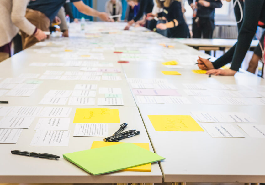 Das Bild zeigt einen langen Tisch, auf dem zahlreiche Zettel und Notizen ausgebreitet sind. Mehrere Personen im Hintergrund sind unscharf dargestellt und scheinen an einem Brainstorming oder einer ähnlichen Gruppenarbeit teilzunehmen, bei der die Zettel auf dem Tisch verwendet werden. Im Vordergrund liegen Stifte und weitere Haftnotizen. Das Bild vermittelt den Eindruck eines intensiven Arbeitsprozesses in einer kollaborativen Umgebung.