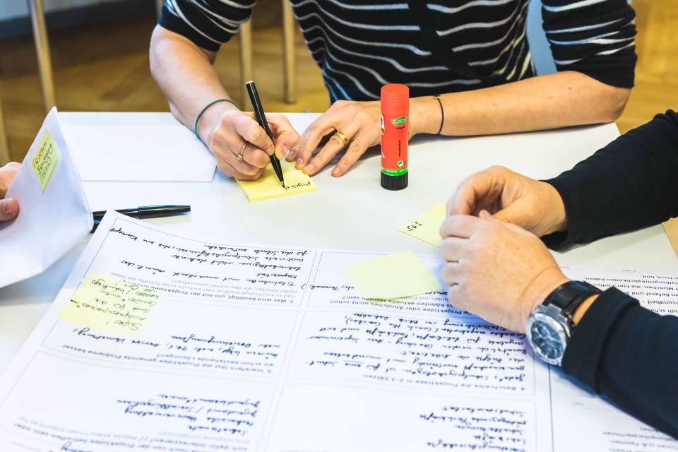 Das Bild zeigt einen Ausschnitt von mehreren Personen, die an einem Tisch sitzen und an einem Projekt arbeiten. Auf dem Tisch liegen zahlreiche Blätter mit handschriftlichen Notizen und Notizzettel, die mit einem Stift beschriftet werden. Es sind Klebstoff und Stifte zu sehen, was auf ein Brainstorming oder eine gemeinschaftliche Planungsarbeit hindeutet. Die Atmosphäre wirkt konzentriert und kollaborativ. Das Bild fokussiert sich auf die Hände und die Schreibutensilien, wobei die Gesichter der Personen nicht sichtbar sind.