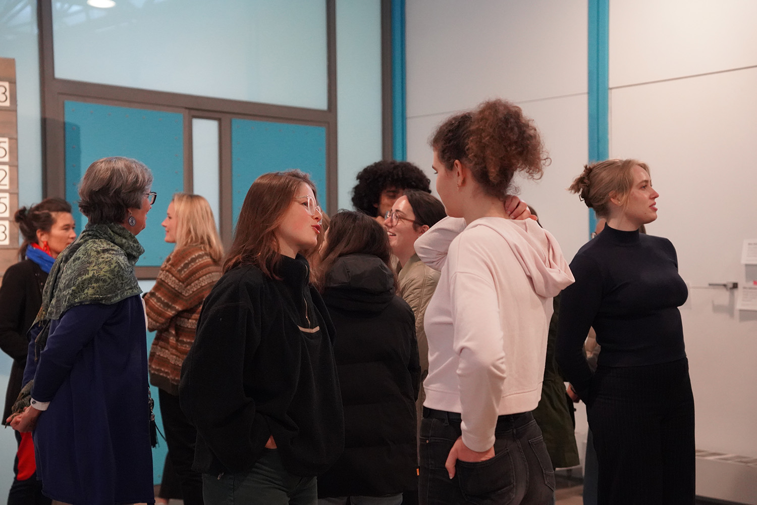Das Bild zeigt eine Gruppe von etwa zehn jungen Erwachsenen und älteren Frauen, die in einem Raum mit hellblauen Wänden und Türen stehen und sich unterhalten. Der Fokus liegt auf zwei jungen Frauen im Vordergrund, die sich intensiv unterhalten. Die anderen Personen im Hintergrund wirken als Teil einer größeren Gruppe, aber ihre Interaktionen sind weniger deutlich. Die Atmosphäre scheint entspannt und ungezwungen zu sein.