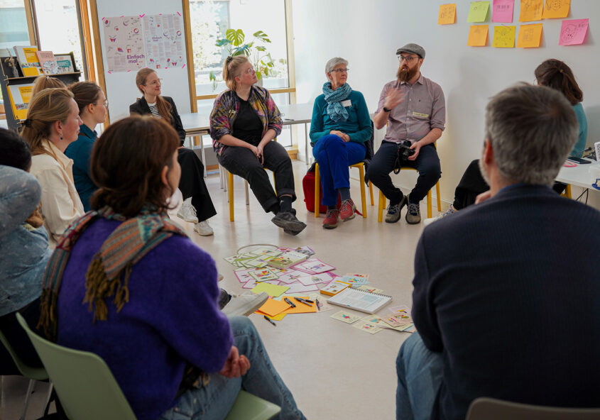 Das Bild zeigt eine Gruppe von Menschen, die in einem hellen Raum in einem Kreis sitzen. In der Mitte des Kreises liegen Karten und Notizbücher auf dem Boden. Ein Mann mit Bart und Mütze gestikuliert und spricht, während die anderen ihm aufmerksam zuhören. An der Wand hängen bunte Zettel mit Zeichnungen. Die Atmosphäre wirkt interaktiv und konzentriert. Das Bild entstand während eines Workshops im Rahmen des Hans Sauer Preis 2025 in dem Münchner Kulturhaus LUISE.