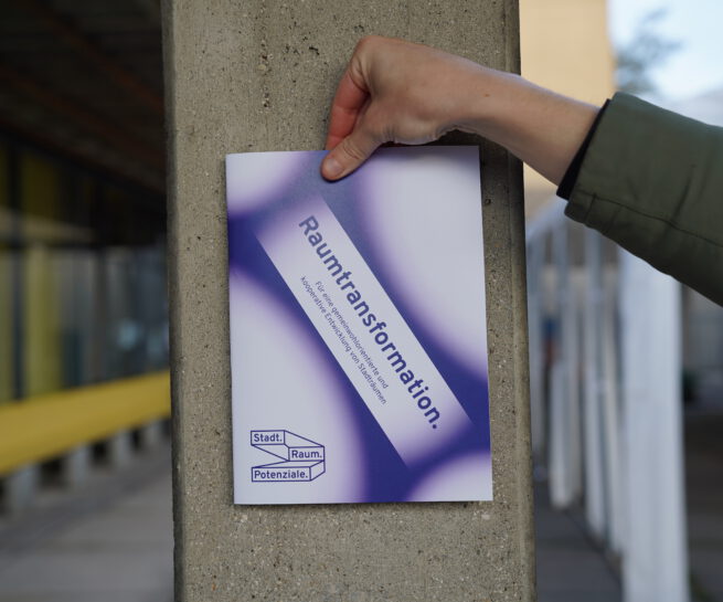 Das Bild zeigt eine Hand, die eine Broschüre mit der Aufschrift "Raumtransformation" gegen eine Betonsäule hält. Im Hintergrund ist der Rest des Gebäudes zu sehen. Mit dem Bild wird die Broschüre präsentiert.