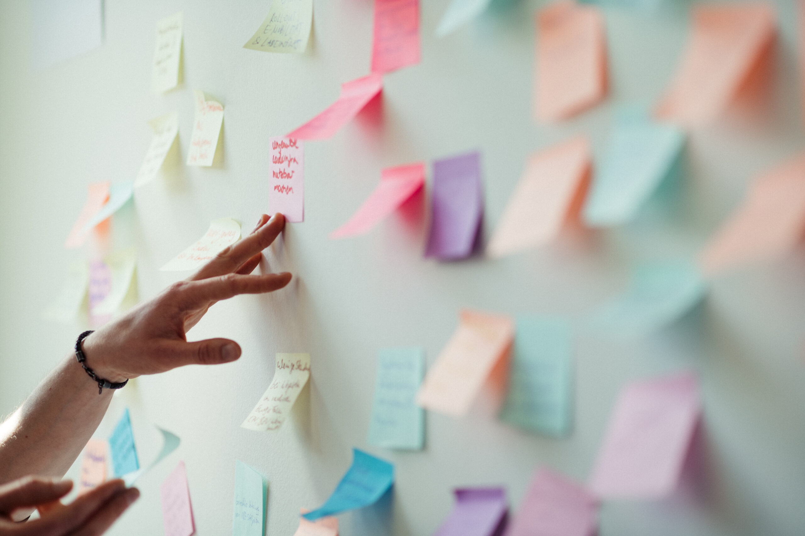 Viele bunte Post-Its kleben an einer Wand. Es ist eine Hand zu sehen, die auf die Post-Its deutet.