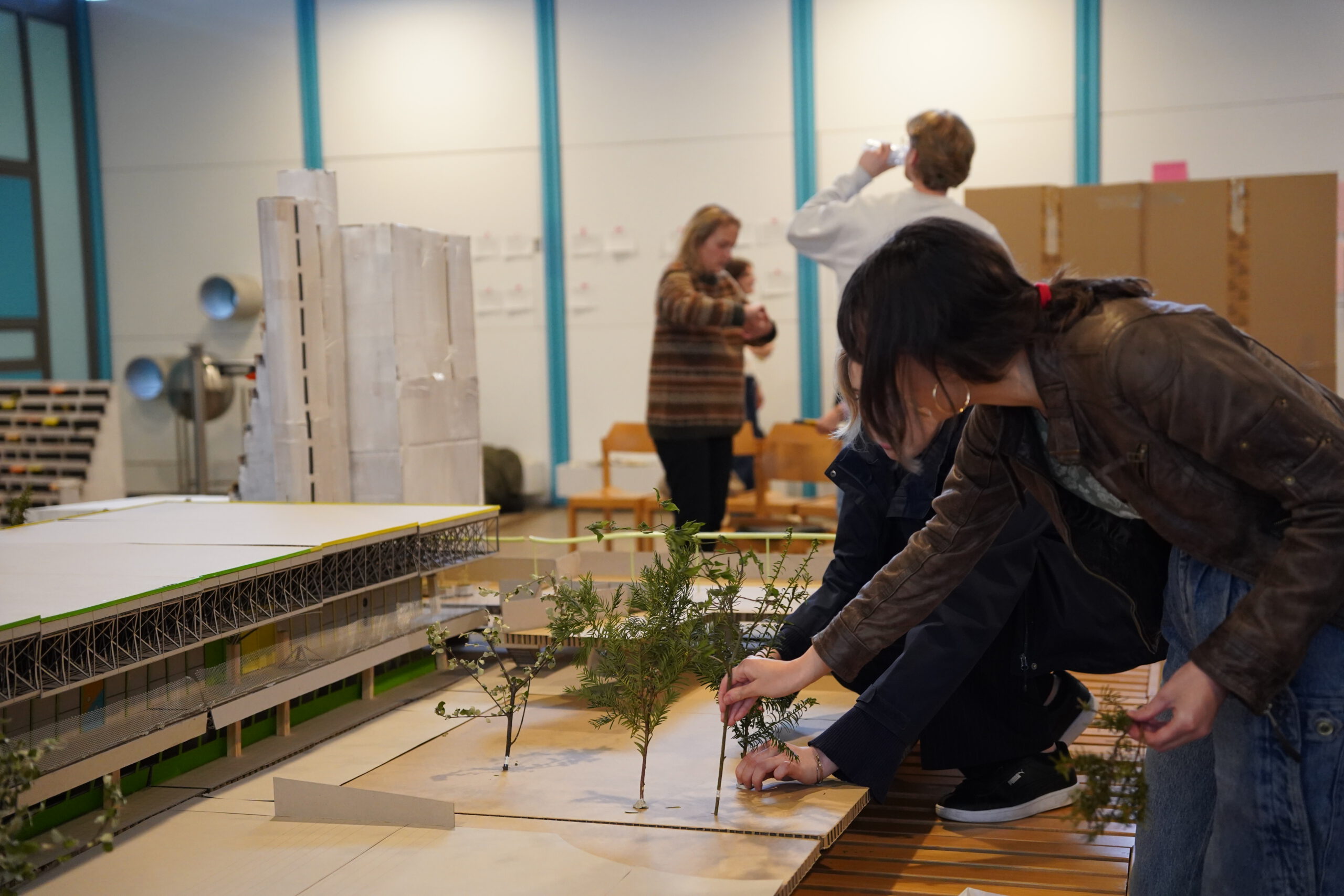 Im Vordergrund sind mehrere Personen zu sehen, die kleine Modelle von Bäumen auf eine Platte setzen. Im Hintergrund sind weitere Menschen zu sehen, eine davon trinkt gerade. Es scheint sich um eine Arbeitssituation zu handeln.