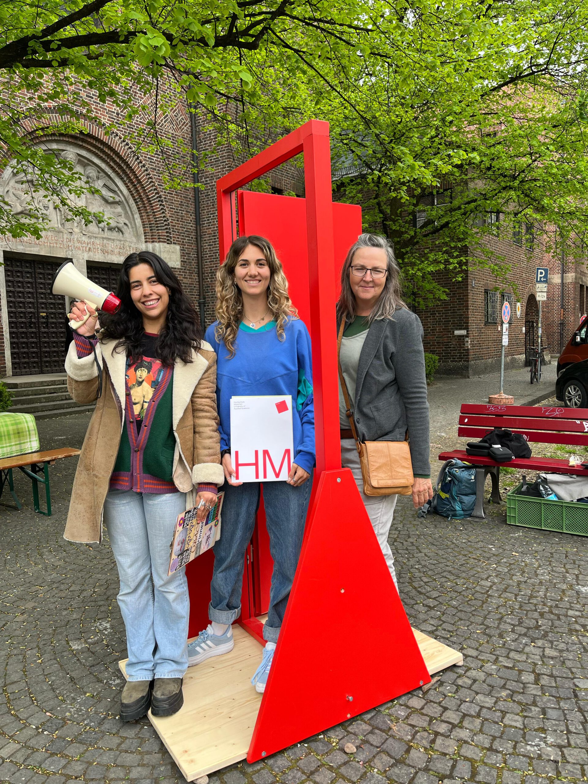 Drei junge Frauen lächeln in die Kamera. Sie stehen in einem Gebilde, das rot angestrichen ist und an eine offene Tür erinnert. Eine Person hält ein Megafon in der Hand, eine weitere eine Broschüre, auf der "HM" steht.