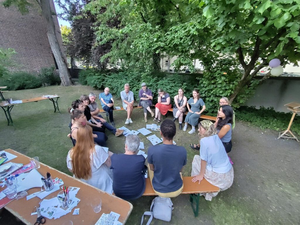 Eine Gruppe von Menschen sitzt in einem Kreis im Garten. In der Mitte sind verschiedene Zettel asgebreitet. Neben der Gruppe steht ein Tisch mit weiteren Zetteln. es scheint sich um eine angeregte Diskussion zu handeln.