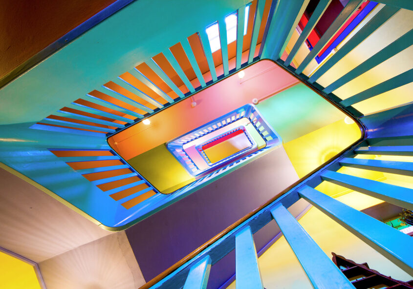Blick nach oben in das farbig gestaltete Treppenhaus des Absalon Gemeinschaftszentrums mit rechteckigem Treppenauge und farbigen Geländern