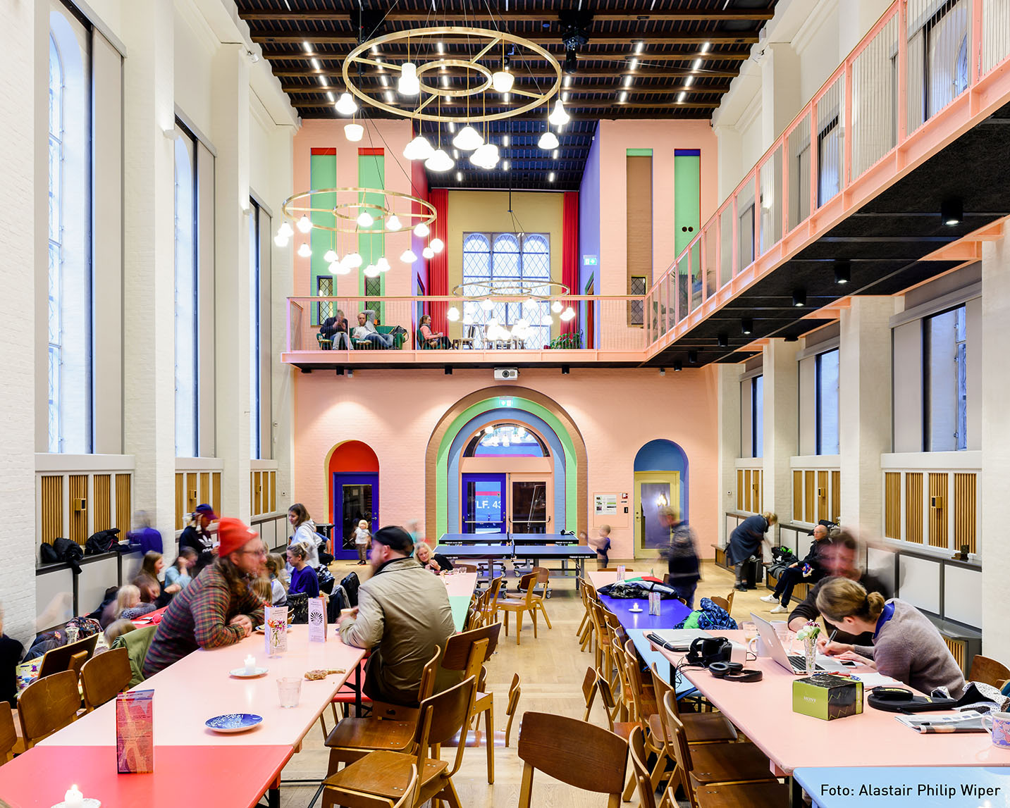 Folkehuset Absalon Main Hall_ Ein farbenfroher Gemeinschaftsraum (die Haupthalle der Folkehuset Absalon Kirche) mit hohen Decken, in dem Menschen an langen Tischen sitzen, mit Balkonen an den Seiten und gewölbten Türen am anderen Ende.