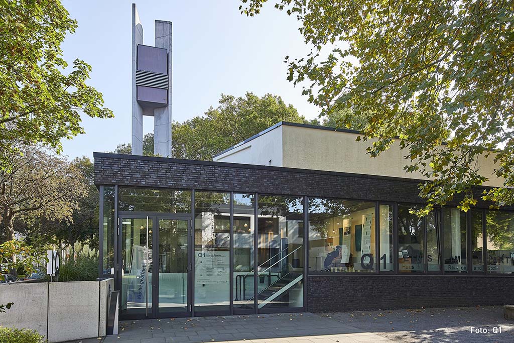 Q1_Bochum Außenansicht des Stadtteilzentrums Q1 in den Räumlichkeiten der Friedenskirche in Bochumg mit großen Glasfronten, dunklem Mauerwerk und einem hohen, weißen Glockenturm im Hintergrund, umgeben von Bäumen.