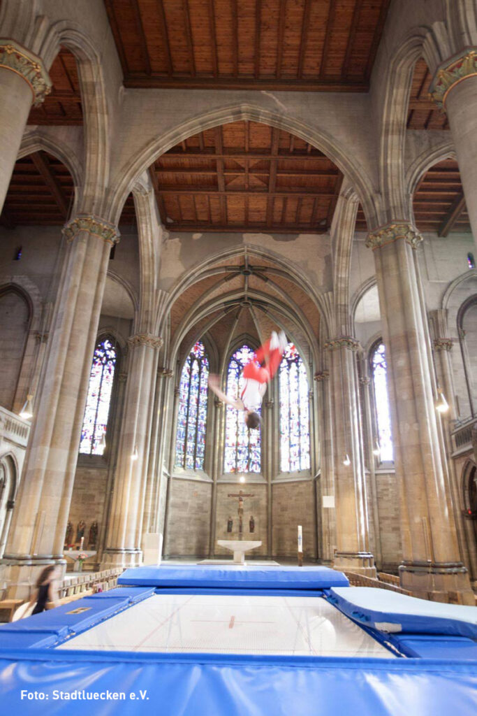 Sankt Maria als.. Eine Person in der Luft vollführt einen Salto auf einem Trampolin in einer großen Kathedrale mit hohen Säulen und Buntglasfenstern.