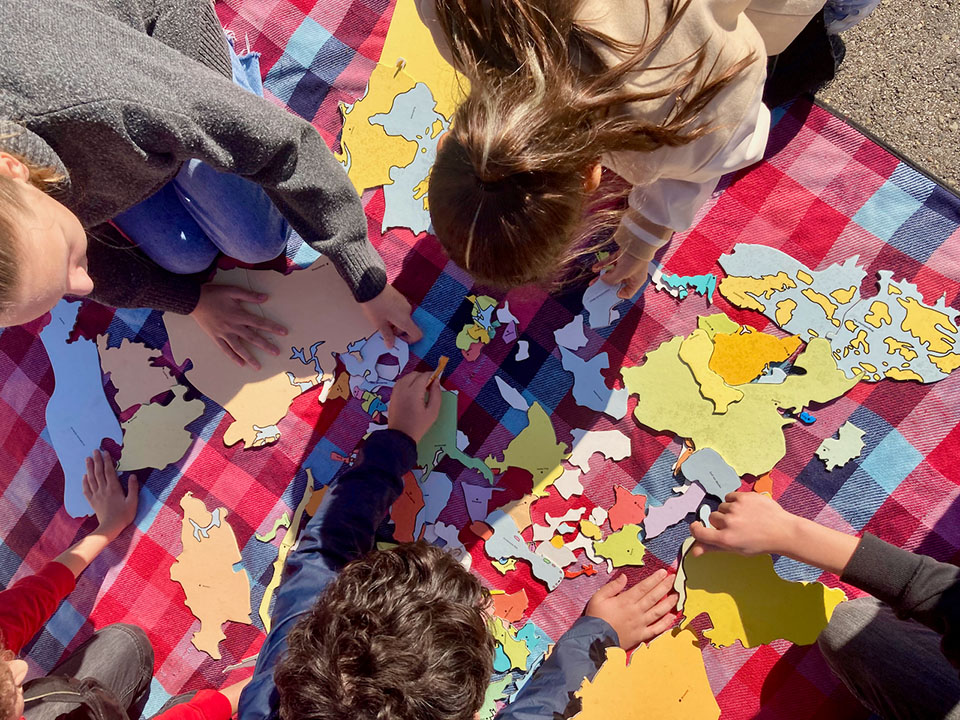 Habibi22_1163 Mehrere Kinder sitzen auf einer karierten Decke und setzen ein großes, buntes Puzzle mit Landkarten zusammen