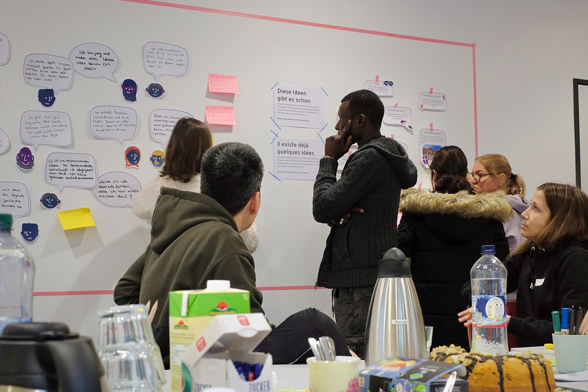 Home not Shelter_Brücken Bauen Gruppe von Menschen steht vor einer Wand mit zahlreichen Zetteln und Sprechblasen, einige sitzen an einem Tisch mit Getränken und Snacks