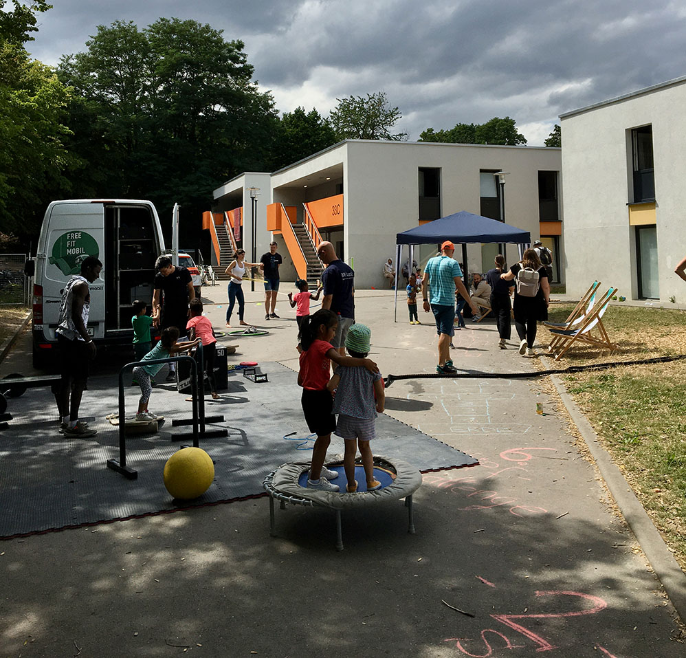 Home not Shelter_Fitness Mobil Kinder spielen auf einem kleinen Trampolin und auf dem Boden vor einem Wohngebäude mit orangenen Treppen, während Erwachsene sich unter einem blauen Pavillon und neben einem weißen Van mit der Aufschrift „FREE FIT MOBIL“ aufhalten