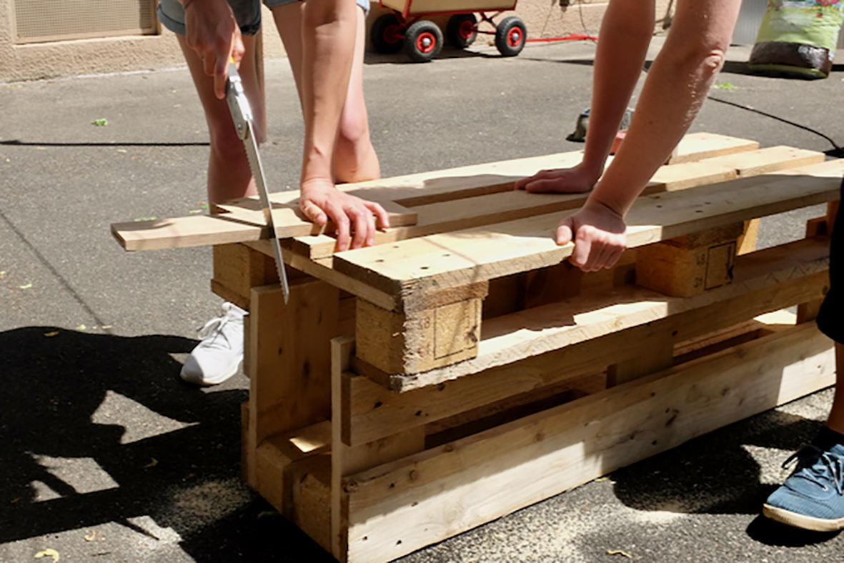 Home not Shelter_Gemeinsam Bänke bauen Zwei Personen sägen und halten Holzbretter auf einem Stapel aus Holzpaletten im Freien
