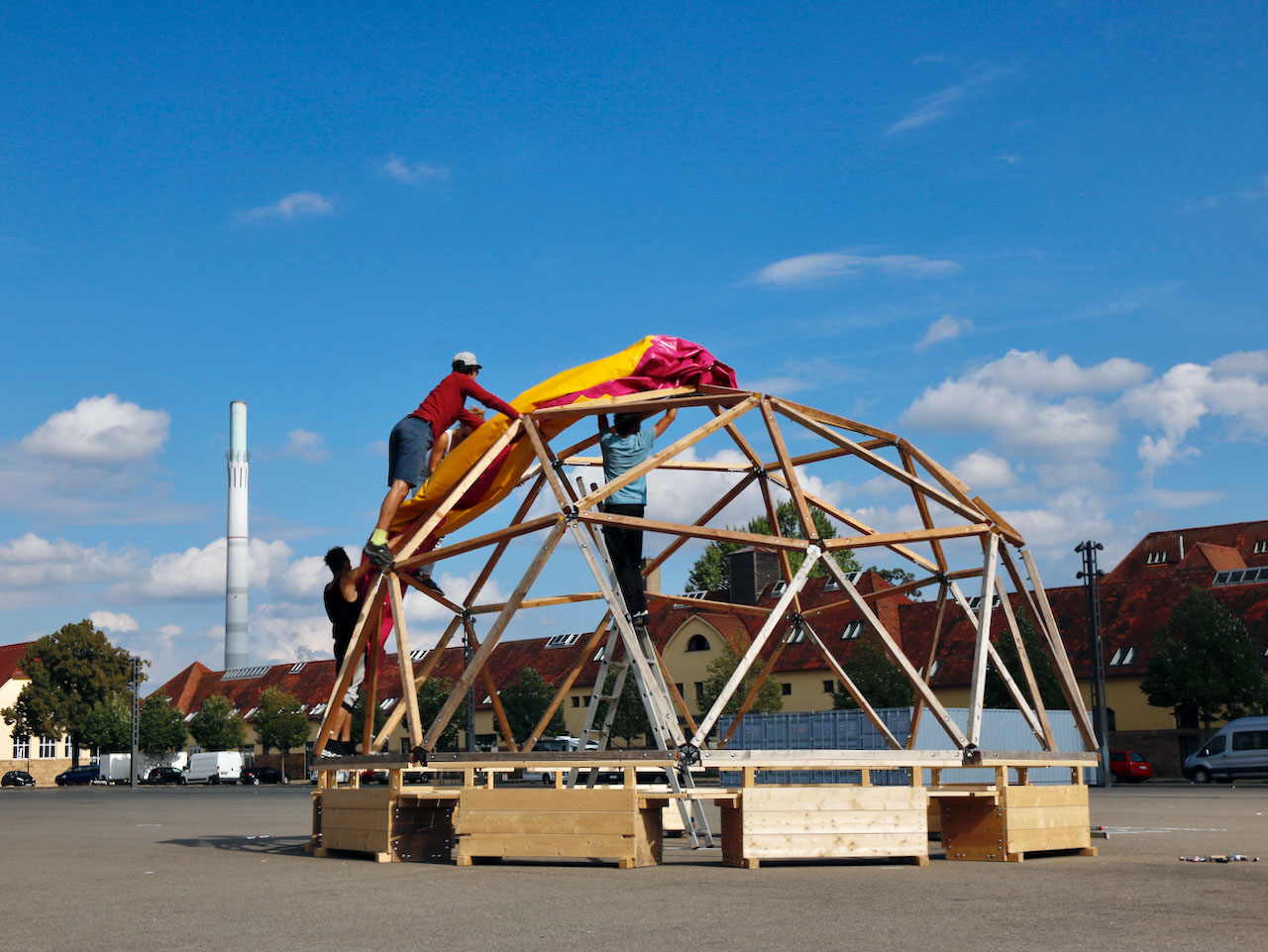 Home not Shelter_Habibi Dome Drei Personen befestigen eine bunte Plane auf einer halbrunden Holzkonstruktion auf einem großen Platz mit Gebäuden im Hintergrund